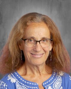 Smiling woman with glasses and long hair, wearing a blue embroidered top, set against a gray background.