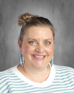 Smiling woman with blue eyes and a bun hairstyle, wearing a striped sweater and feather earrings, against a gray background.