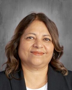 A smiling woman with wavy brown hair, wearing a black blazer, against a gray background.