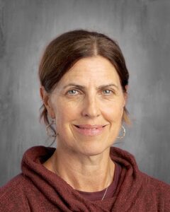 A woman with brown hair and blue eyes smiles, wearing a burgundy hoodie and hoop earrings, against a gray background.