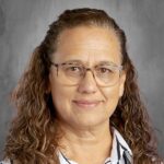 A woman with curly hair and glasses smiles, wearing a striped shirt against a grey background.