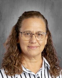 A woman with curly hair and glasses smiles, wearing a striped shirt against a grey background.