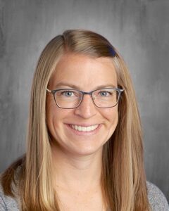 Smiling woman with long, straight blonde hair and glasses, wearing a gray sweater against a neutral background.