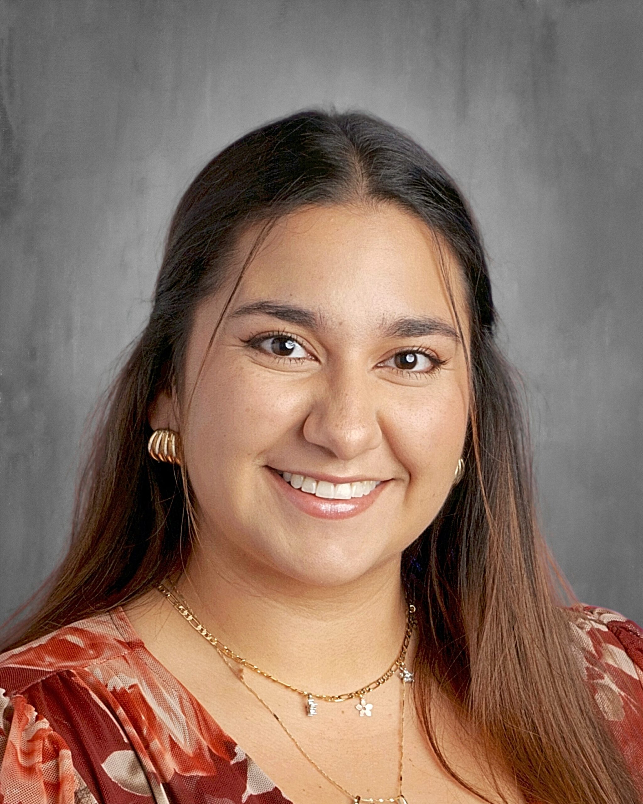 A smiling woman with long brown hair, wearing floral attire and layered necklaces, poses against a gray background.