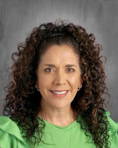 A smiling woman with curly hair, wearing a green top, poses against a gray background.