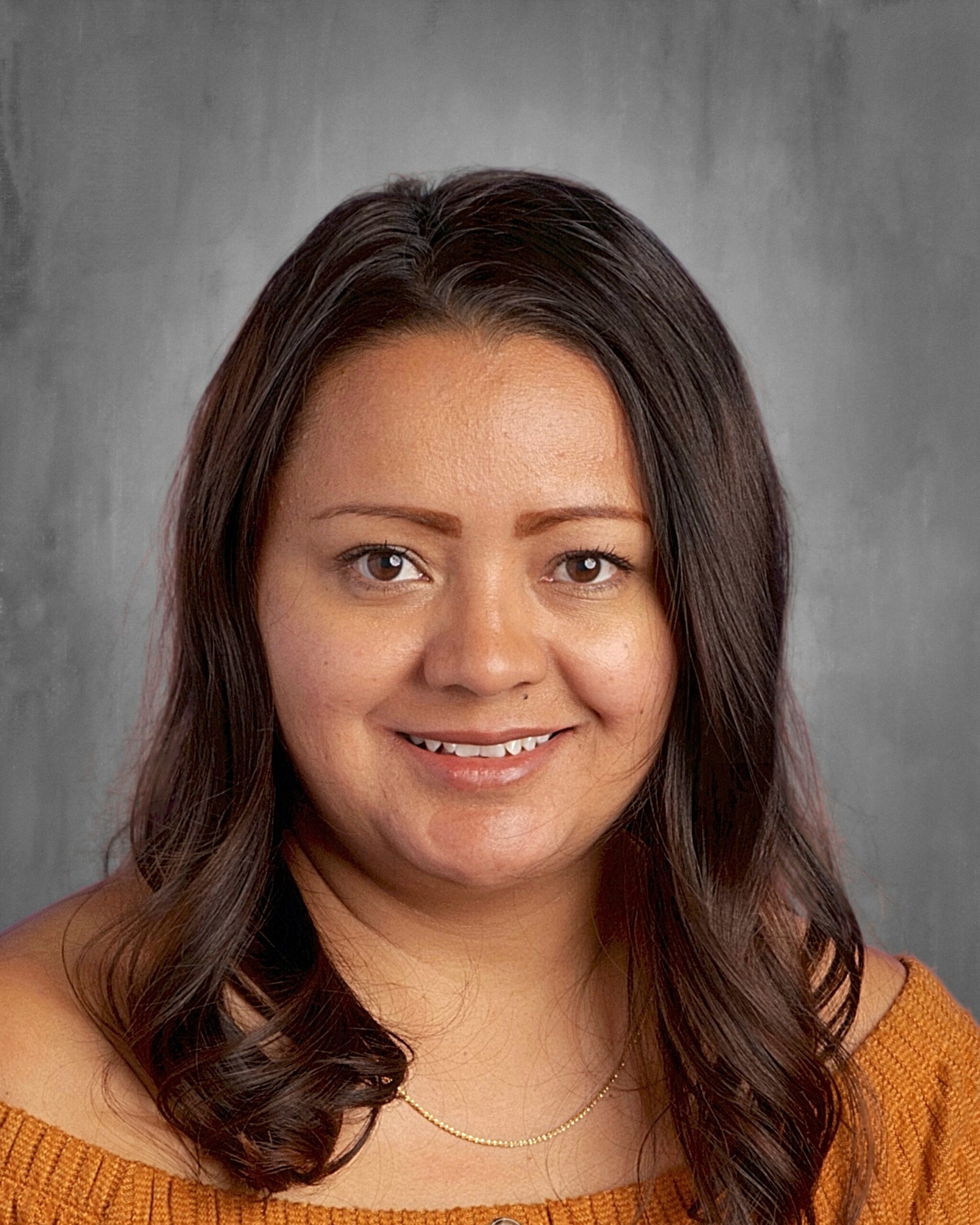 Smiling woman with long dark hair, wearing an orange off-shoulder top, against a gray background.