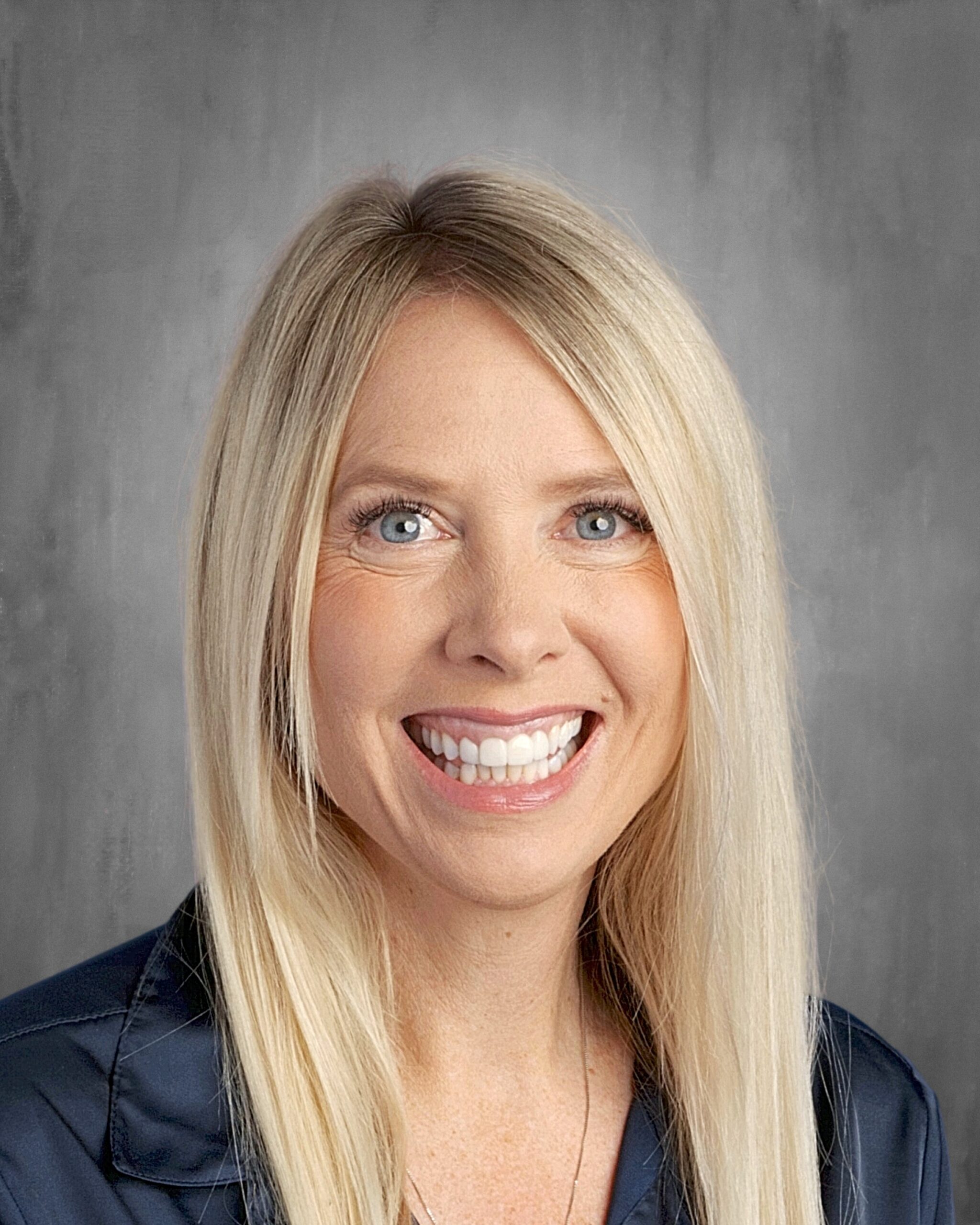 A smiling woman with long blonde hair and blue eyes, wearing a dark shirt, against a gray background.