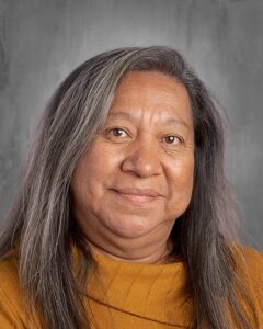A woman with long, straight gray hair smiles, wearing a mustard-colored top against a gray background.