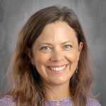 A smiling woman with long brown hair and blue eyes, wearing a purple knit top, against a gray background.
