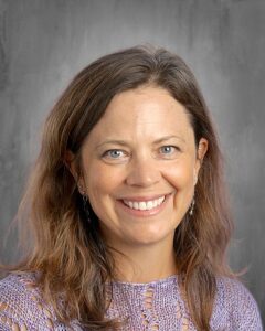 A smiling woman with long brown hair and blue eyes, wearing a purple knit top, against a gray background.
