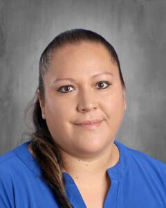 A woman with long dark hair, wearing a blue shirt, smiles against a gray background.