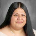 Portrait of a young woman with long black hair, wearing a light sweater and a silver necklace, smiling gently.
