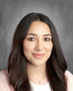 A young woman with long, dark hair smiles, wearing a light pink sweater and a delicate necklace, against a gray background.