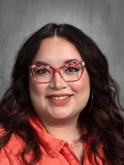 A smiling woman with long dark hair, wearing colorful glasses and an orange shirt, poses for a portrait against a gray background.